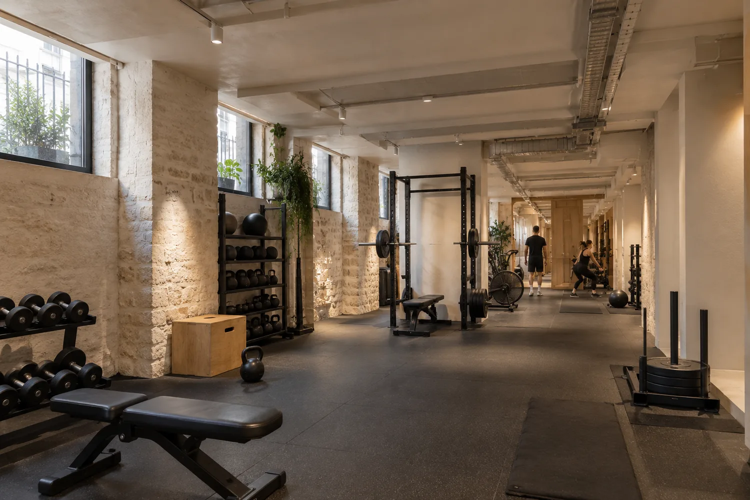 Salle de sport aménagée dans un volume industriel en pierre, racks de musculation et haltères sous poutres apparentes