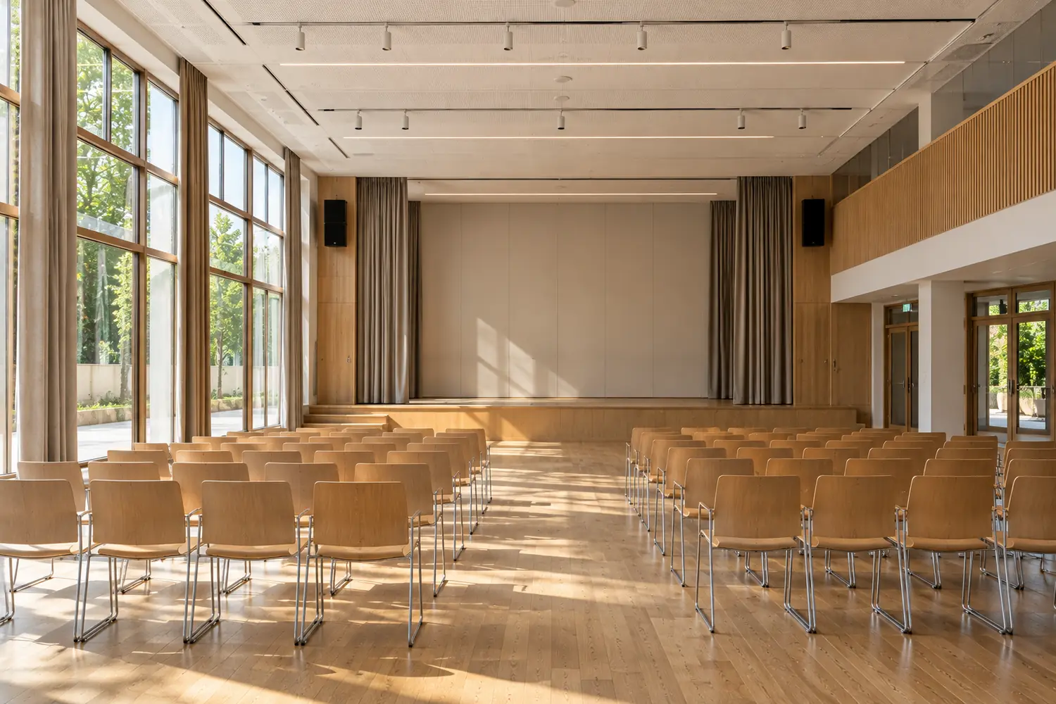 Salle polyvalente vide en rez-de-chaussée, rangées de chaises face à une estrade basse, grandes baies latérales, vue frontale, sans texte lisible