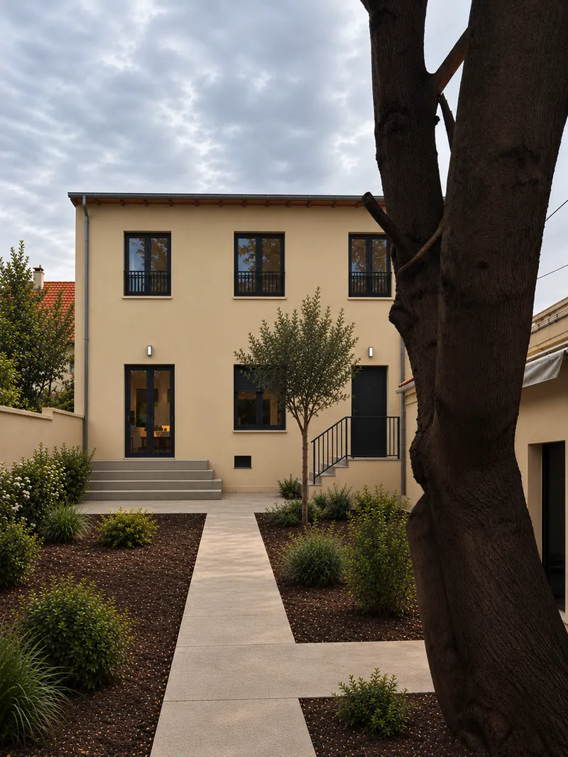 Même maison après surélévation : un étage de trois fenêtres à garde-corps sur une façade enduite beige, menuiseries noires, jardin planté, grand arbre à droite.