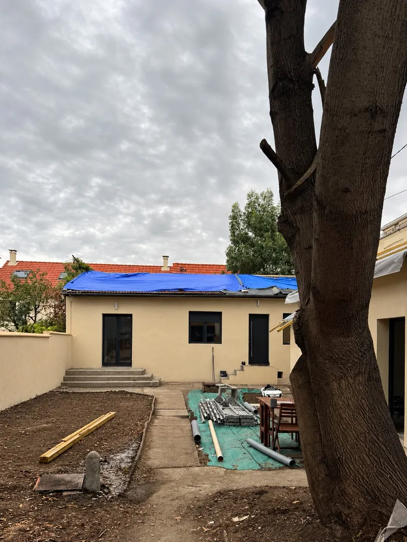 Maison de plain-pied avant travaux : façade enduite beige, toiture recouverte d’une bâche bleue, jardin en chantier, grand arbre à droite.