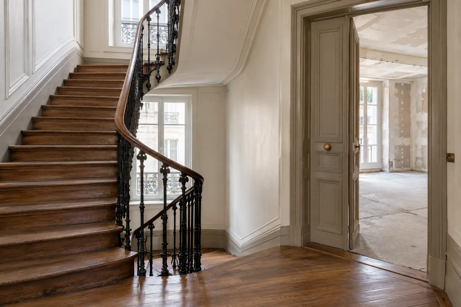 Cage d’escalier d’un immeuble parisien ancien, rampe en fer forgé, porte palière ouverte sur un appartement en travaux, sans personnage